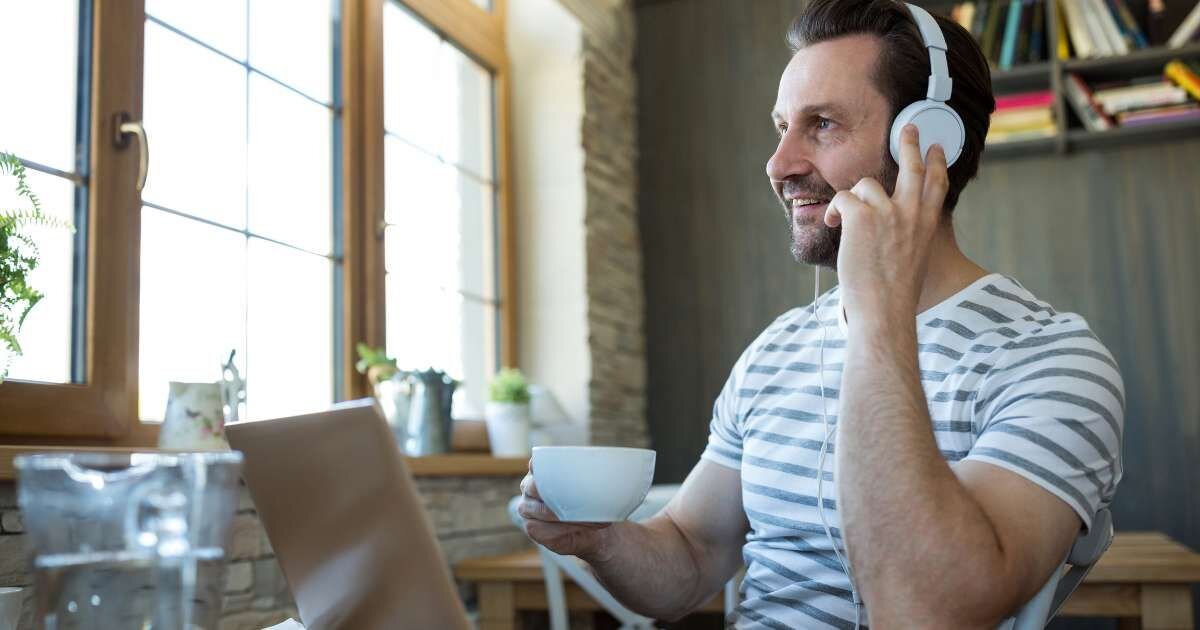 man wearing headphones, holding is coffee, while sitting at his desk 